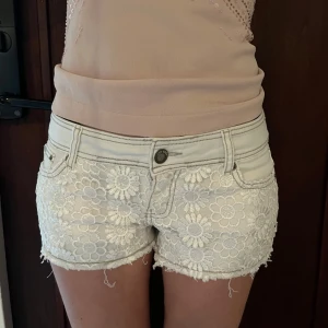 Vita  jeansshorts - Supersöta vita lågmidjade jeansshorts med blommig spets framtill och på ena bakfickan 🩷Midja : 37 cm 