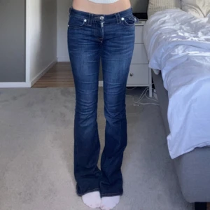 True Religion bootcut jeans blå - Snygga blå bootcut jeans från True Religion med låg midja och klassiska kontrastsömmar. Jeansen har dekorativa fickor bak med lock och knappar samt detaljerade sömmar framtill.