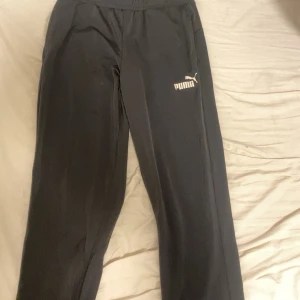 Svarta träningsbyxor från Puma M - Svarta träningsbyxor från Puma med resår i midjan och snörning. Byxorna har en rak passform och Puma-logga på låret. Perfekta för träning eller chill hemma. Materialet känns lätt och smidigt.