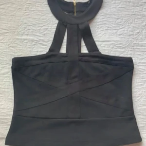 Svart halterneck korsett med dragkedja - Snygg svart korsettopp med halterneck och bred krage. Toppen har coola banddetaljer framtill och en guldig dragkedja bak som ger en edgy vibe. Perfekt för dig som vill sticka ut med en stilren och modern look. Storlek lapp saknas. Hade på mig en gång.