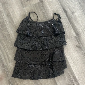 Svart paljettklänning med volanger - Supersnygg svart kortklänning med tunna axelband och flera lager volanger. Hela toppen är täckt av glittriga paljetter som ger en festlig look. Perfekt för dig som vill sticka ut och glänsa på dansgolvet eller andra tillfällen