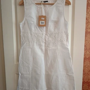 Vit kort klänning med spetsdetaljer - Superfin vit kort klänning med spetsdetaljer i midjan och längst ner. Ärmlös modell med rund halsringning, perfekt för varma dagar. Klänningen har en enkel och fräsch look som passar till många tillfällen.