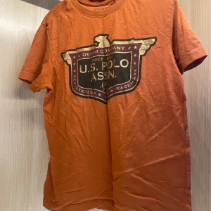Orange t-shirt från U.S. Polo Assn. - Snygg orange t-shirt från U.S. Polo Assn. med stort tryck framtill i svart och guld. Klassisk rund halsringning och korta ärmar. Tillverkad i mjuk bomull, perfekt för en avslappnad och cool stil. Stl m men passar också S