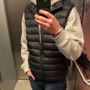 Svart dunväst  - Säljer en svart dunväst med quiltad design och dragkedja framtill. Västen har en klassisk, sportig look och passar perfekt över en hoodie eller tröja. Den är lätt och smidig att bära, perfekt för kyliga dagar.