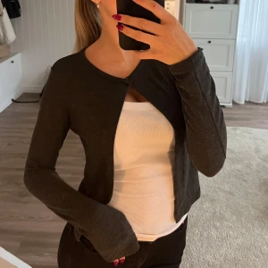 Mörkgrå croppad kofta från Gina Tricot - Säljer en mörkgrå croppad kofta från Gina Tricot. Koftan har lång ärm, rund halsringning och knäpps med en knapp framtill. Perfekt att slänga över ett linne eller topp för en enkel och stilren look.