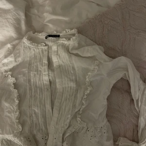 Vit blus med spets och volang från Zara - Supersöt vit blus från Zara med knappar framtill, spetsdetaljer och volanger längs bröstet. Blusen har broderade detaljer
