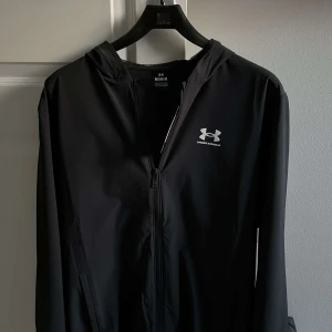 Svart vindjacka från Under Armour - Säljer en svart vindjacka från Under Armour med huva och dragkedja framtill. Jackan har en stilren design med logga på bröstet och är perfekt för träning eller vardag. Lätt och smidig att bära med sig.