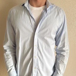 Ljusblå skjorta från Oscar Jacobson - Säljer en klassisk ljusblå skjorta från Oscar Jacobson med knappar framtill. Skjortan har lång ärm och är tillverkad i ett mjukt bomullstyg från Albini. Perfekt för en stilren och clean look.