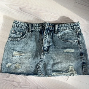 Blå jeanskjol med slitningar - Säljer en blå jeanskjol i klassisk denim med slitna detaljer framtill och baktill. Kjolen har fyra fickor både fram och bak. Säljer för 300, men kan gå ner i pris vid köp direkt. Kjolen är helt ny och ej använd!