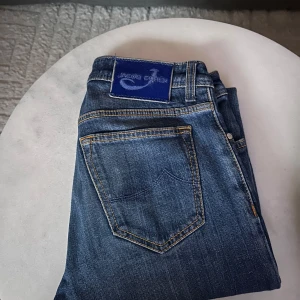 Blå jeans från Jacob Cohën - Säljer ett par klassiska blå jeans från Jacob Cohen med raka ben och snygga kontrastsömmar. Jeansen har fem fickor, knappgylf och en mörkblå patch med logga bak i midjan. Perfekta för dig som gillar stilrena och tidlösa jeans. Hör av er vid minsta fundering! 