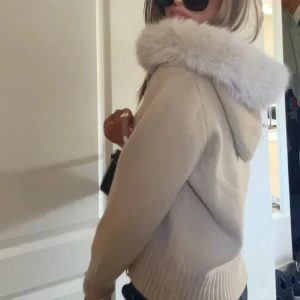 Beige stickad alessa jacka  - Mysig beige stickad jacka med huva och bred päls runt huvkanten. Jackan har dragkedja framtill och ribbade muddar vid ärmslut och nederkant. Perfekt för kyliga dagar och ger en trendig look.
