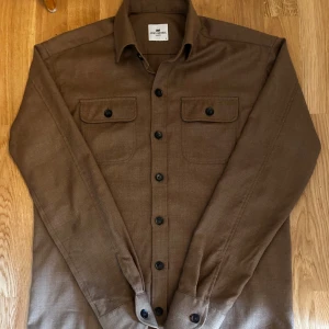 Brun overshirt från John Henric  - En brun overshirt från John Henric som knappast är använd och ej har några defekter. Perfekt nu till lite kallare sommarkvällar. Tveka inte på att höra av er vid minsta fråga samt om ni vill ha fler bilder. Pris går att diskutera!