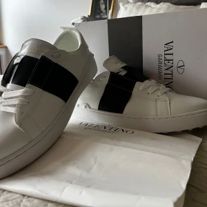 Valentino Garavani - Snygga vita sneakers från Valentino Garavani med en bred svart rem över mitten och ikoniska nitar på hälen. Skorna har rund tå, platt sula och klassisk snörning. Perfekta för dig som gillar stilrena och exklusiva sneakers.