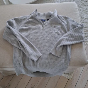 Ljusgrå stickad tröja med dragkedja från GANT - Säljer en ljusgrå stickad tröja från GANT med halv dragkedja och broderad logga på bröstet. Tröjan har lång ärm och ribbad struktur, perfekt för kyligare dagar. Klassisk och stilren modell.