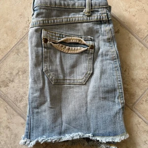 Ljusblå jeanskjol från MIMI - Lågmidjad ljusblå jeanskjol från MIMI med råa, fransiga kanter och slitna detaljer. Klassisk femficksmodell med bälteshällor och knappgylf. Snygga bakfickor med dekorativa sömmar. Perfekt för sommaren!