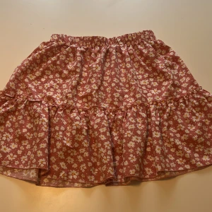 Rosa blommig kjol från SHEIN - Supersöt ljusröd kjol från SHEIN med vitt blommönster och volang nertill. Kjolen har resår i midjan och är perfekt för en somrig look. Lätt och luftig modell som är enkel att matcha med olika toppar.