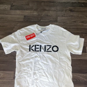 Vit t-shirt  - Säljer en vit t-shirt med stor svart logga framtill. Klassisk rund halsringning och normal passform. Perfekt för dig som gillar stilrena och trendiga plagg med tydlig branding.