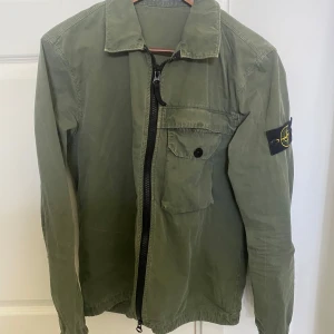 Grön overshirt från Stone Island - Säljer en grön overshirt från Stone Island med klassisk patch på ärmen. Jackan har dragkedja framtill, bröstficka med knapp och en avslappnad passform. Perfekt för lager-på-lager och snygg till jeans.