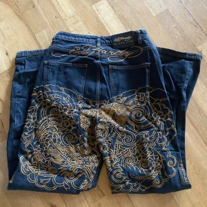 Mörkgråa Ed Hardy jeans med broderi - Mörk gråa baggy jeans från Ed Hardy Black Line med stora, detaljerade guldiga broderier på bakfickorna och Ed Hardy-logga broderad ovanför fickan. Kan användas för både kvinnor och män. Köptes för 800kr och säljer för 750kr!