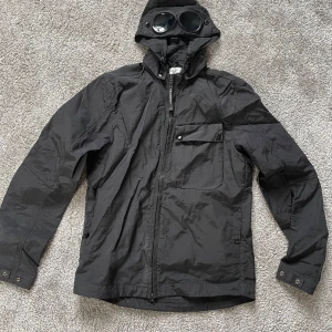 Svart Goggle Jacket från C.P. Company - Säljer en svart Goggle Jacket från C.P. Company med huva och ikoniska glasögon i huvan. Jackan har dragkedja framtill, bröstficka med knapp och är tillverkad i lättviktig nylon. Perfekt för dig som gillar streetwear och unika detaljer.