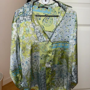 Mönstrad skjorta från ZARA - Säljer en färgglad skjorta från ZARA i storlek S. Skjortan har ett patchwork-liknande blommigt och paisleymönster i grönt, blått och vitt. Långärmad med klassisk krage och knäppning framtill. Perfekt för dig som gillar unika plagg! Använd endast en gång. Superfin till sommaren!