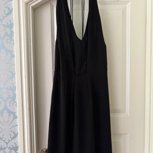 Svart långklänning med djup rygg och tunna axelband - Säljer en elegant svart långklänning med djup v-ringning och öppen rygg. Klänningen har tunna axelband som knäppsi nacken och ger en stilren siluett. Perfekt för dig som vill ha en minimalistisk och tidlös look.