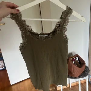 Olivgrön linnetopp med spets - Supersöt olivgrön linnetopp med breda axelband och fina spetsdetaljer längs ringningen och axlarna. Perfekt att styla till jeans eller kjol för en trendig look.