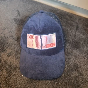 Marinblå keps med 500-lapp patch - Snygg marinblå keps från Bruce Lee x All Cap One med mesh på sidorna och bak. Framtill sitter en patch som föreställer en svensk 500-kronorssedel. Justerbar rem baktill för perfekt passform.