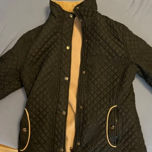 Quiltad jacka  - Super söt  svart quiltad jacka med dekorativa sömmar och gulddetaljer vid fickorna. Jackan har tryckknappar, dragkedja och lång ärm. Perfekt för dig som gillar stilren design med lite extra detaljer. M, funkar för S också. 