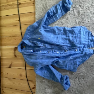 Blå linneskjorta från Polo Ralph Lauren - Säljer en blå långärmad skjorta i linne från Polo Ralph Lauren. Skjortan har klassisk krage, knappar framtill och den ikoniska gula broderade loggan på bröstet. Perfekt för en stilren och avslappnad look.