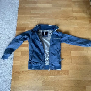 Blå vindjacka från Sail racing - Säljer en blå vindjacka från Salracing med dragkedja. Jackan har ribbade muddar och två fickor med dragkedja. Perfekt för blåsiga dagar och enkel att matcha med jeans.