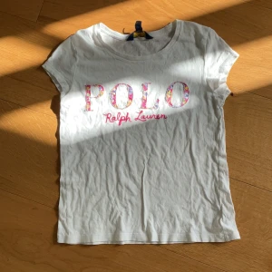 Vit t-shirt från Polo Ralph Lauren - Vit t-shirt från Polo Ralph Lauren med färgglad blommig text på bröstet och rosa broderad logga. Klassisk rund halsringning och korta ärmar. Perfekt för en avslappnad och stilren look.