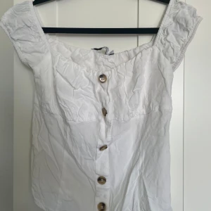 Vit blus med knappar - Säljer en söt vit blus med korta ärmar och knappar framtill. Blusen har en lätt rynkad kant upptill och är tillverkad i ett tunt bomullstyg. Perfekt för varma dagar och enkel att matcha med jeans eller kjol.