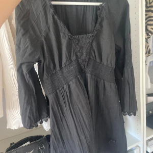 Svart klänning med v-ringning och smock - Svart klänning med v-ringad hals och smock i midjan. Långa ärmar med spetsdetaljer vid ärmslut. Klänningen har en lätt rynkad kjol och dekorativa sömmar framtill. Perfekt för dig som gillar en bohemisk stil.