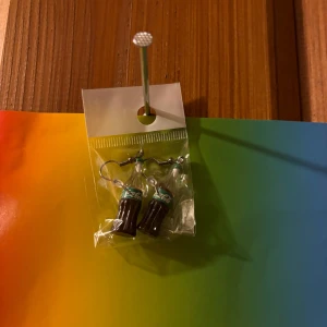 Örhängen med colaflaskor - Säljer ett par unika örhängen formade som små colaflaskor. Perfekt för dig som vill ha något annorlunda och lekfullt i öronen. Färgerna är brunt och silver med detaljer i turkos på flaskans etikett.