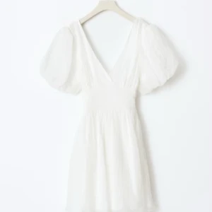 Vit kortklänning med puffärmar och rosett (bow dress) - Superfin vit kortklänning med v-ringning, puffiga kortärmar och stor rosett i ryggen. Klänningen har en omlottkänsla och markerad midja, perfekt för sommarens alla tillfällen. Tillverkad i ett lätt, luftigt material som ger en romantisk vibe. Perfekt till studenten eller bara en nice sommardag. Helt ny och oanvänd, prislapp hänger på, snygg men slutsåld på hemsida.