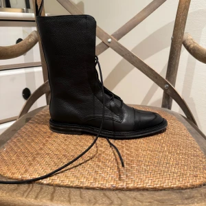 Svarta läderboots från INCH2 - Säljer ett par svarta boots i äkta läder från INCH2. Skorna har snörning framtill, rund tå och en stilren, hög siluett. Perfekta för dig som gillar minimalistisk och edgy stil. Kommer i originalkartong.