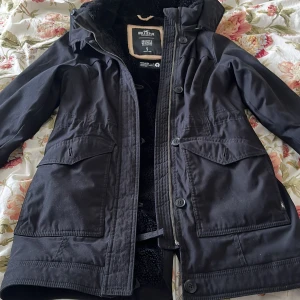 Svart parkajacka från Hollister - Svart parkajacka från Hollister med huva och dragsko i midjan. Jackan har stora fickor framtill, stängs med dragkedja och knappar, och är fodrad med mjuk pälsimitation. Perfekt för kyliga dagar.