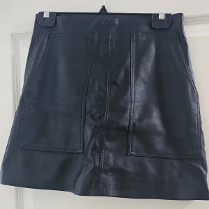 Svart skinnkjol från H&M - Säljer en svart skinnkjol från H&M i storlek 36. Kjolen är kort med två stora fickor framtill och har en stilren, minimalistisk design. Perfekt för dig som gillar en edgy och trendig look.