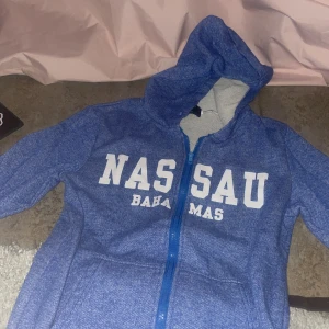 Blå hoodie från Bahamas Shirt Co. - Blå hoodie med vit text 'NASSAU BAHAMAS' på bröstet. Tröjan har dragkedja framtill och huva. Perfekt för en avslappnad stil och enkel att matcha med jeans eller mjukisbyxor.