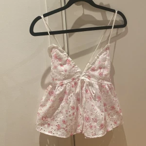 Vit blommig linnetopp - Söt vit linnetopp med rosa blommigt mönster och broderade detaljer. Toppen har v-ringning, knyt framtill och är ärmlös. 