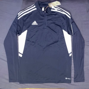 Blå långärmad träningströja från Adidas - Säljer en blå långärmad träningströja från Adidas med vita detaljer och dragkedja vid halsen. Tröjan har klassiska Adidas-ränder på axlarna och är tillverkad i ett syntetmaterial som passar perfekt för sport och träning.