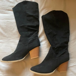 Svarta boots med klack - Säljer ett par svarta boots med hög skaft och bred träfärgad klack. Skorna har rund tå och är tillverkade i mocka-liknande material. Oanvända 