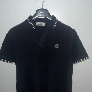 Mörkblå  piké t-shirt från Stone Island - Säljer en mörkblå piké t-shirt från Stone Island med klassisk krage och knappar framtill. T-shirten har korta ärmar med vita ränder på krage och ärmslut samt en broderad logotyp på bröstet.
