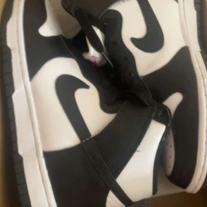 Nike Dunk high svartvita sneakers - Säljer ett par Nike Dunk Low sneakers i svart och vitt. Skorna har klassisk låg modell med svart swoosh, svarta snören och vit tå med perforeringar. Yttermaterialet är syntet och sulan är platt och svart. Perfekta för en stilren streetlook.