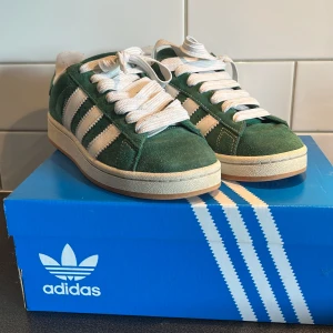 Adidas Campus 00s gröna sneakers - Säljer ett par Adidas Campus 00s sneakers i mörkgrön mocka med vita ränder och vit sula. Klassisk retrostil med rund tå och snörning. Perfekta för dig som gillar streetwear och vill sticka ut med en snygg färg på fötterna. Passar både killar och tjejer! Perfekt nu till sommaren, ett par gröna snören medföljer också. Skriv vid funderingar! Priset är inte hugget i sten, och kan diskuteras.