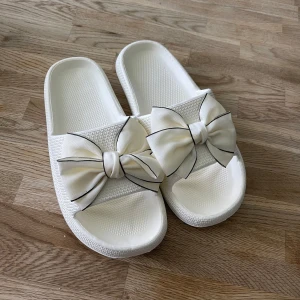 Vita sandaler med rosett - Säljer ett par vita tofflor med stora dekorativa rosetter framtill. Skorna har en platt sula och är tillverkade i syntetmaterial. Perfekta för sommaren och ger en söt touch till din outfit.Helt nya,EU storlek 39