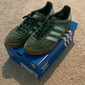 Adidas Gazelle Indoor gröna sneakers - Säljer ett par gröna Adidas Gazelle Indoor sneakers med klassiska ljus blå  ränder och gummisula. Skorna har snörning och är tillverkade i mocka med detaljer i syntet. 