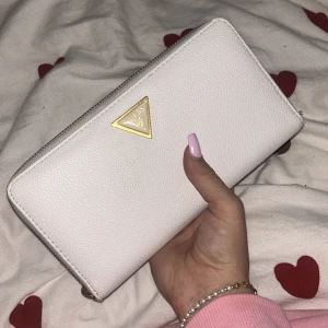 Vit plånbok från Guess med gulddetaljer - Säljer en stilren vit plånbok från Guess med guldfärgad dragkedja och logga. Plånboken har flera kortfack, myntfack med dragkedja och plats för sedlar. Insidan är metallic guld. Perfekt för att hålla ordning på dina kort och pengar.