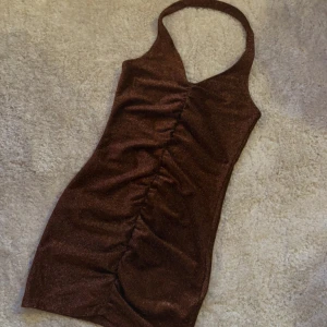 Brun glittrig halterneckklänning - Säljer en brun glittrig kortklänning med halterneck och rynkade detaljer framtill. Klänningen har en tight passform och är perfekt för dig som vill sticka ut. Supersnygg till fest eller utekväll! Helt ny och endast testad